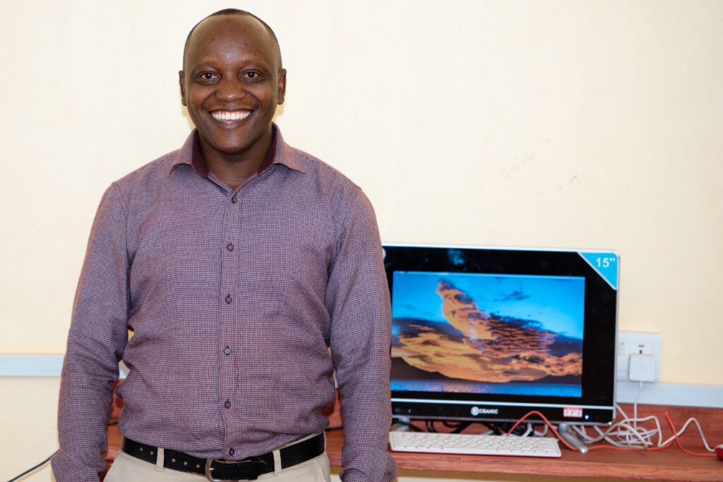 Teacher and computer manager Eligi Tairo smiles next to a computer in the classroom