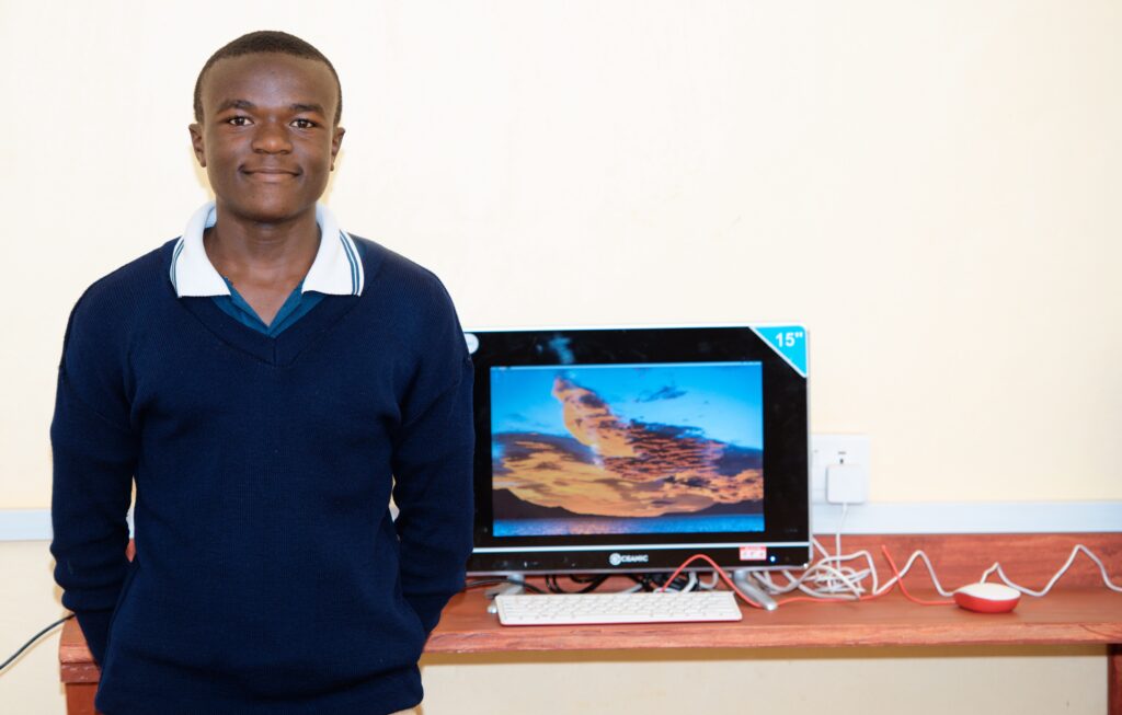 Student Larson smiles standing next to a computer in his classroom